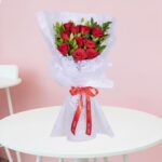 A bouquet of vibrant red roses and fresh green leaves arranged and wrapped in non-woven paper, finished with a bright red satin ribbon.